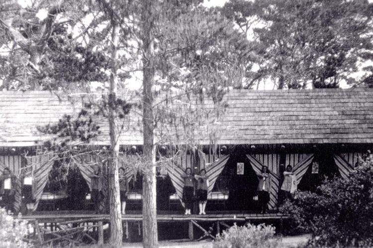 Historic black and white photo of women standing outside rustic tent cabins beneath tall trees at Asilomar.