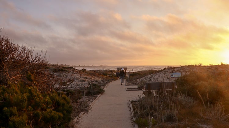 A couple walks along a wooden boardwalk leading through sandy dunes toward the sea at sunset.